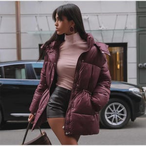 May include: A burgundy puffer jacket with a drawstring waist and a hood. The jacket is worn over a light pink turtleneck and black shorts. A brown handbag is held in the hand. The background includes a car and a building.