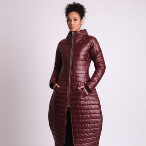 May include: A long, burgundy puffer coat with a high collar and a full-length zipper. The coat has a unique, rounded shape at the bottom and features pockets. The model is wearing black pants and black platform boots. The coat is designed for cold weather.