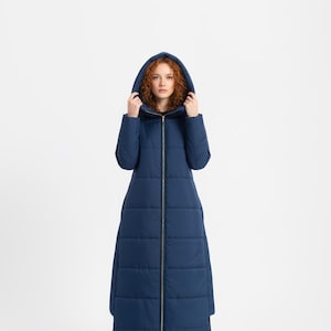 Può includere: Lungo cappotto trapuntato blu navy con cappuccio e cerniera dorata a tutta lunghezza. Il cappotto è lungo fino al pavimento e presenta un design trapuntato. La persona indossa stivali neri.