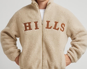 May include: A beige fleece jacket with a high collar and a full zipper. The word "HILLS" is embroidered in brown block letters across the chest. The model is wearing brown corduroy pants.