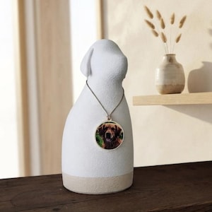 Puede incluir: Una figura con forma de perro, blanca y texturizada, con una base lisa de color claro. Un cordón fino rodea el cuello de la figura y sostiene un colgante circular que muestra una fotografía en color de un perro marrón con la boca abierta.