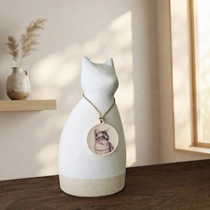 Puede incluir: Una escultura de cerámica blanca texturizada con forma de gato y base beige. Un cordón fino rodea el cuello y sostiene un colgante circular con una ilustración realista y detallada de un gato atigrado gris y blanco mirando hacia la derecha.