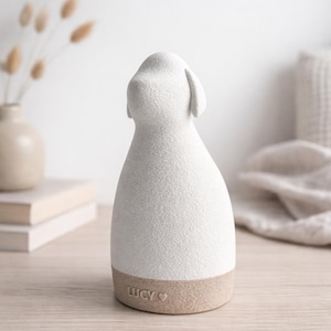 Puede incluir: Figura de perro de cerámica blanca con superficie texturizada y base beige. La figura tiene un diseño minimalista y la palabra "LUCY" está escrita en la base. El perro está de espaldas al espectador.