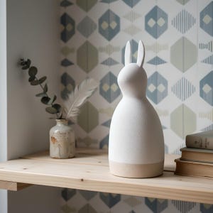 May include: A white ceramic rabbit figurine with a smooth, matte finish and a rounded, tapered body. The sculpture features long, upright ears and a flat base, standing as a decorative object alongside a small ceramic vase and stacked books.