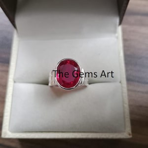 May include: A silver ring with a large, oval-cut ruby gemstone. The ruby is a vibrant red color and is set in a simple, elegant band. The ring is displayed in a white jewelry box. The text "The Gems Art" is visible.