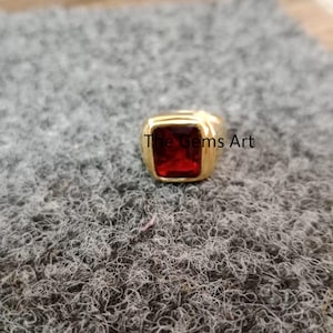May include: A gold-colored ring with a large, square, red gemstone. The ring is set on a textured, gray surface. The text "The Gems Art" is visible on the ring.