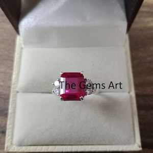 Könnte beinhalten: Ein silberner Ring mit einem großen, rechteckigen, leuchtend roten Edelstein als Mittelpunkt. Kleinere, klare Edelsteine flankieren den Hauptstein. Der Ring wird in einer weißen Schmuckschatulle präsentiert. Der Text "The Gems Art" ist sichtbar.