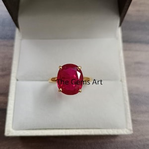 May include: A gold ring with a large, oval-shaped ruby gemstone. The ruby is a deep red hue and is held in place by four prongs. The ring is presented in a white jewellery box. Text on the ring says "The Gems Art".