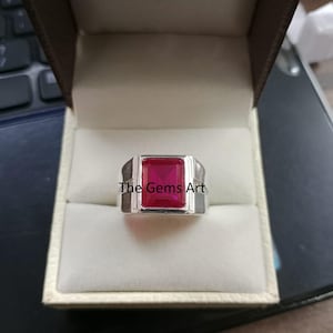 May include: A silver ring featuring a large, square, vibrant red gemstone. The ring has a simple, contemporary design and is presented in a jewellery box. The text "The Gems Art" is visible on the ring.