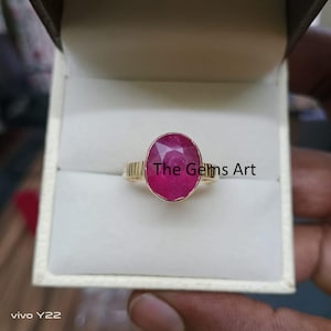 Op de afbeelding: Een gouden ring met een grote, ovaalvormige, dieprode edelsteen. De ring heeft een gouden band met een textuur. De tekst "The Gems Art" is zichtbaar op de edelsteen.