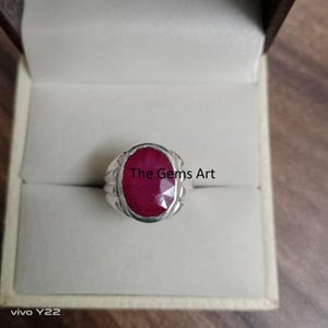 Może przedstawiać: Srebrny pierścionek z dużym, owalnym, ciemnoczerwonym kamieniem szlachetnym. Pierścionek ma prosty, klasyczny design i jest prezentowany w jasnym pudełku. Na kamieniu widoczny jest napis "The Gems Art".