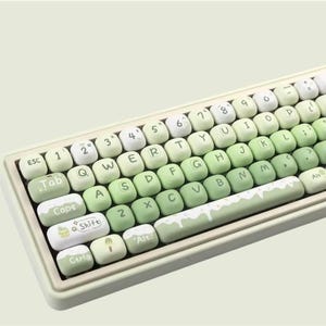 Peut inclure: Un clavier vert pastel et blanc avec des touches arrondies. Les touches sont disposées selon une disposition QWERTY standard, avec des chiffres et des lettres. Le clavier a un design décoratif sur la barre d'espace et certaines touches.