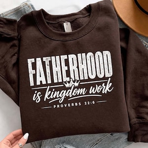 Op de afbeelding: Een donkerbruine sweatshirt met de witte tekst "FATHERHOOD is kingdom work" en een kleine kroongrafiek. De tekst "PROVERBS 22:6" staat eronder gedrukt. De sweatshirt ligt op een blauwe jeans.