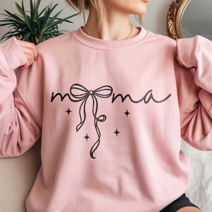 Puede incluir: Sudadera rosa claro con la palabra "mama" en cursiva, adornada con un lazo y cuatro estrellas. La parte superior de manga larga está hecha de un material suave.