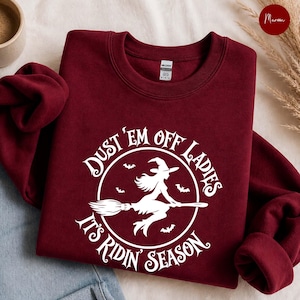 May include: A maroon crewneck sweatshirt featuring a white graphic of a witch flying on a broomstick inside a circular frame. The text reads DUST 'EM OFF LADIES IT'S RIDIN' SEASON, accompanied by small bat silhouettes surrounding the central witch figure.