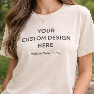 May include: Cream-colored t-shirt with the text "YOUR CUSTOM DESIGN HERE" in dark gray, along with "Made to Order for You" below. The shirt is short-sleeved and appears to be made of a soft fabric. The model is wearing a gold necklace.