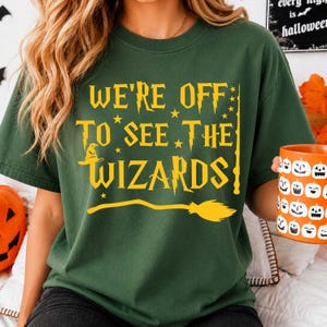 May include: A dark green t-shirt featuring yellow text that reads WE'RE OFF TO SEE THE WIZARDS. The design includes a small sorting hat icon, several stars, a vertical wand graphic, and a horizontal broomstick illustration across the bottom.