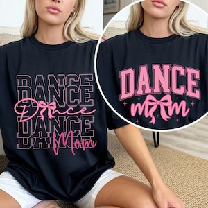 May include: Black t-shirt with the words "DANCE Mom" in pink block letters and a pink bow graphic. The word "DANCE" is stacked multiple times in a bold font. The shirt is a casual style.