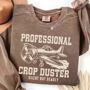May include: Brown sweatshirt with the words "PROFESSIONAL CROP DUSTER" and an illustration of a crop duster airplane. The phrase "SILENT BUT DEADLY" is printed below the airplane graphic. A silver necklace with a pendant is visible.