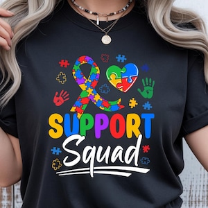 Op de afbeelding: Zwart t-shirt met een kleurrijk grafisch ontwerp. Het ontwerp bevat een puzzellint, een puzzelhart, handafdrukken en de woorden "SUPPORT Squad" in levendige kleuren. Het shirt heeft een ronde hals.