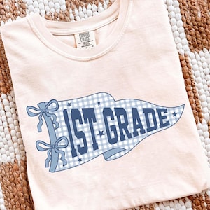 May include: A light peach-colored t-shirt featuring a graphic of a blue and white gingham pennant flag. The flag displays the text 1ST GRADE in bold blue letters, accented with small blue stars and two blue ribbons tied on the left side.