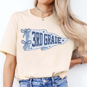 Könnte beinhalten: Ein cremefarbenes Kurzarm-T-Shirt mit einer blau-weiß karierten Wimpelgrafik. Der Wimpel enthält den Text 3RD GRADE in fetten blauen Buchstaben, akzentuiert durch zwei blaue Schleifen auf der linken Seite und kleine blaue Sternformen.