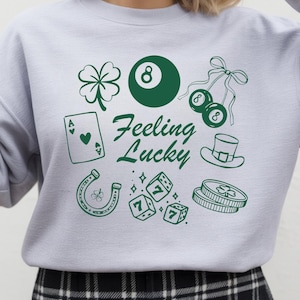 Op de afbeelding: Een lichtgrijs sweatshirt met een groen grafisch ontwerp met een klavertjevier, een achtbal, dobbelstenen en de tekst "Feeling Lucky". Andere afbeeldingen zijn een hoefijzer, een speelkaart en munten.