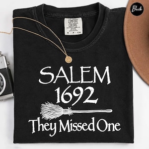 Op de afbeelding: Een zwart t-shirt met korte mouwen en witte tekst met de tekst SALEM 1692 They Missed One, met een afbeelding van een bezemsteel tussen de datum en de laatste zin. Het shirt heeft een Comfort Colors merklabel bij de nek en een ronde hals.