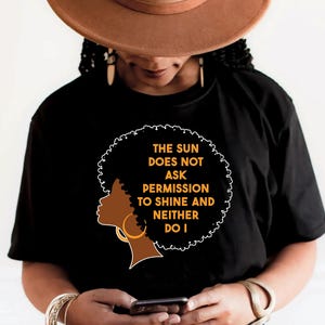 Op de afbeelding: Zwart T-shirt met een afbeelding van een vrouwensilhouet met gouden oorringen en een afro. De tekst luidt: "DE ZON VRAAGT GEEN TOESTEMMING OM TE SCHIJNEN EN IK OOK NIET."