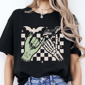 May include: A black t-shirt featuring a graphic print of a green monster hand and a white skeleton hand performing a pinky promise. The design includes a beige and black checkered background, two flying bats, and small four-pointed star accents.