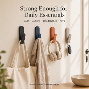 Puede incluir: Cuatro ganchos de pared en negro, azul, naranja y gris, cada uno sosteniendo una bolsa de mano, una chaqueta, auriculares y llaves. El texto dice "Strong Enough for Daily Essentials" y "Hand-finished 3D printed design."