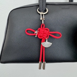 May include: A black leather handbag with a red decorative knot charm. The charm features a silver-colored fan pendant and a small letter charm. The bag has a structured shape and a top handle.