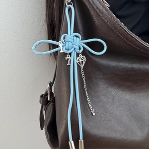 May include: A light blue knotted charm with silver accents, including the number 7 and a diamond-shaped pendant, hangs from a brown leather bag. The charm is attached with a silver clasp.