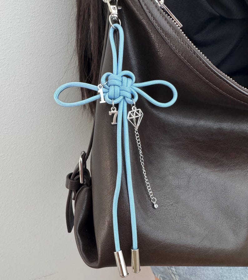 May include: A light blue knotted charm with silver accents, including the number 7 and a diamond-shaped pendant, hangs from a brown leather bag. The charm is attached with a silver clasp.