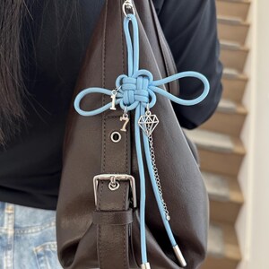 May include: A brown leather handbag with a silver buckle and a light blue cord accent. The cord is tied in a decorative knot and features charms with the numbers "1" and "7", and a diamond shape.