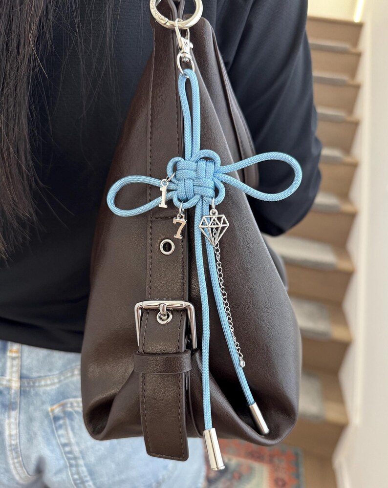 May include: A brown leather handbag with a silver buckle and a light blue cord accent. The cord is tied in a decorative knot and features charms with the numbers "1" and "7", and a diamond shape.