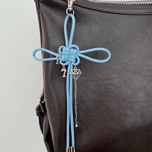 May include: A light blue knotted cord charm with silver-colored charms, including a key, the number 7, and a diamond shape, hangs from a silver clasp. The charm is attached to a dark brown leather bag.