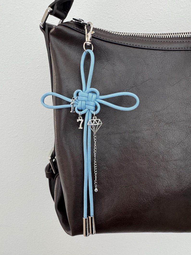 May include: A light blue knotted cord charm with silver-colored charms, including a key, the number 7, and a diamond shape, hangs from a silver clasp. The charm is attached to a dark brown leather bag.