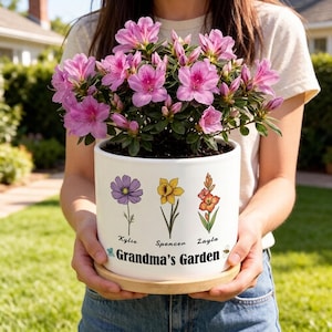 May include: A white ceramic flower pot with a floral design and the text "Grandma's Garden" holds a blooming pink azalea plant. The pot features illustrations of purple, yellow, and orange flowers with names below. The pot sits on a wooden saucer.