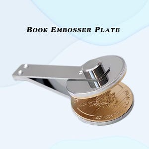 May include: A silver book embosser plate with a circular die and a lever arm. The die is pressed onto a gold-colored coin, creating an embossed design. The text "Book Embosser Plate" is displayed above the embosser.