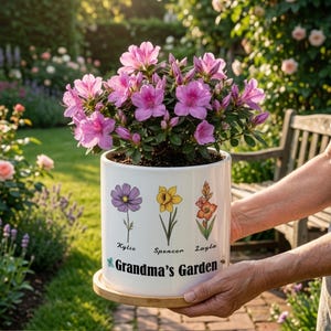 May include: A white ceramic flower pot with a floral design and the text "Grandma's Garden." The pot holds a vibrant pink flowering plant. The pot has a wooden base and is held by a person.
