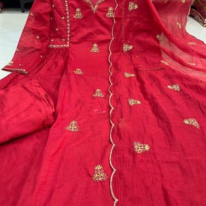 Pode incluir: Um vestido vermelho vibrante com intrincados bordados dourados. A peça de manga comprida apresenta uma borda ondulada e delicados enfeites ao redor do decote e das mangas. O vestido é feito de um tecido fluido e inclui calças combinando.