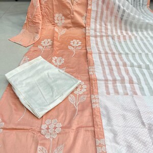 K&ouml;nnte beinhalten: Pfirsichfarbenes, lang&auml;rmliges Tunika- und Hosen-Set mit wei&szlig;er Blumenstickerei. Ein gestreifter wei&szlig;er und pfirsichfarbener Schal ist seitlich drapiert. Das Outfit ist traditionell.