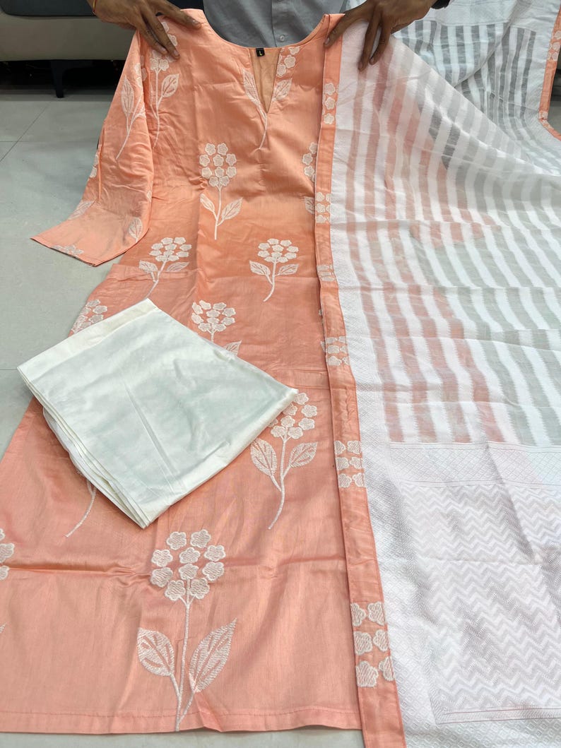 K&ouml;nnte beinhalten: Pfirsichfarbenes, lang&auml;rmliges Tunika- und Hosen-Set mit wei&szlig;er Blumenstickerei. Ein gestreifter wei&szlig;er und pfirsichfarbener Schal ist seitlich drapiert. Das Outfit ist traditionell.