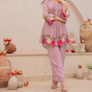 May include: A two-piece lavender outfit featuring a tunic and pants. The tunic has a pleated bodice, three-quarter sleeves with floral embroidery, and a scalloped hem. The pants are a matching lavender color.