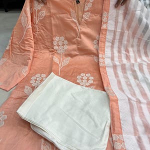 K&ouml;nnte beinhalten: Pfirsichfarbenes, besticktes Kurta-Set mit wei&szlig;en floralen Details. Das Set beinhaltet ein lang&auml;rmliges Oberteil, eine wei&szlig;e Hose und einen gestreiften Schal. Das Kurta hat einen V-Ausschnitt und ist mit wei&szlig;en Blumenstickereien verziert.