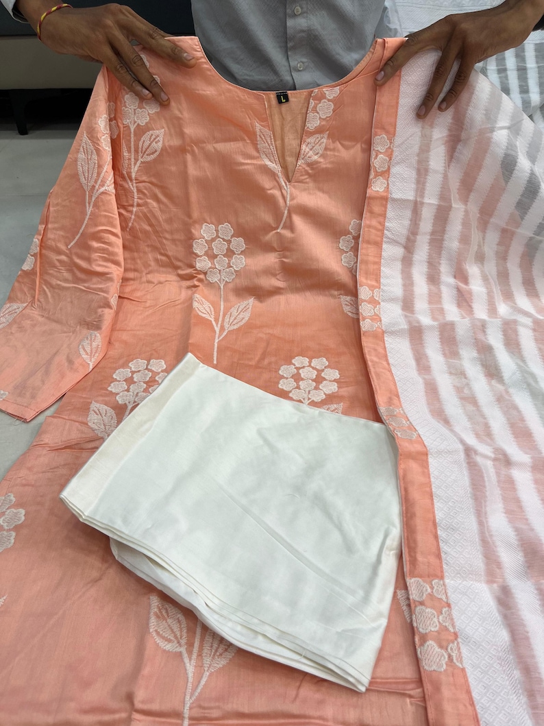 K&ouml;nnte beinhalten: Pfirsichfarbenes, besticktes Kurta-Set mit wei&szlig;en floralen Details. Das Set beinhaltet ein lang&auml;rmliges Oberteil, eine wei&szlig;e Hose und einen gestreiften Schal. Das Kurta hat einen V-Ausschnitt und ist mit wei&szlig;en Blumenstickereien verziert.