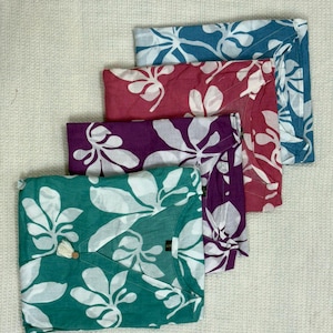 May include: Four folded tops in teal, magenta, purple, and blue, each with a white floral pattern. The teal top has a tassel and button detail at the neckline. The tops are arranged on a white textured surface.