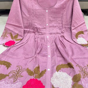 May include: A light purple tunic shirt with a round neckline and long sleeves. The shirt has vertical pleats down the front and is decorated with embroidered floral designs in white, brown, and pink along the hem and sleeve cuffs.