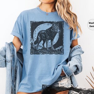 May include: Light blue t-shirt with a black and white wolf illustration. The wolf is standing in front of a full moon. The shirt is made of a soft material and has a relaxed fit. The words "Washed Denim" are printed on the shirt.